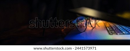 Glasses lie on the laptop, reflecting light from the screen in the dark.