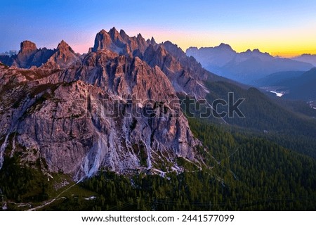 Aerial view from high altitude of Dolomite peaks lit by pink light of  the sunset sky, with Lago di Misurina valley and Dolomites mountain panorama against blue sky with a yellow horizon band. Italy Royalty-Free Stock Photo #2441577099