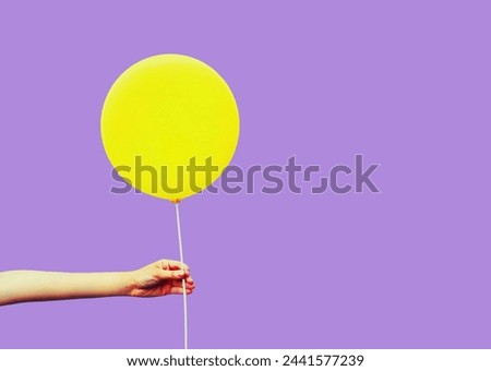 Colorful image close up female hand holding or showing yellow balloon on violet background