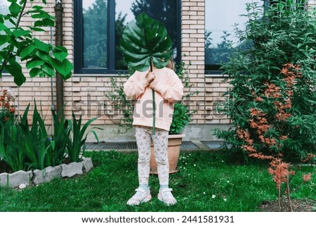 Smiling little girl holding monstera leaf in front of house in green garden. Child carrying big monstera leaf outdoors. Concept of Ecology, Consumerism, Individuality, Sustainability, Idea.