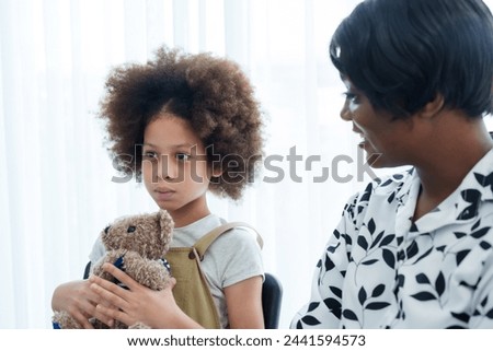 Young child with curly hair clutches teddy bear, wary glance at camera, mother beside provides comfort during clinic visit. Little one with voluminous curls holds stuffed toy, apprehension in eyes,
