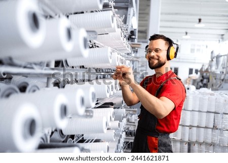 Textile industry. Factory worker with earmuffs standing by large industrial knitting machine and checking threads. Royalty-Free Stock Photo #2441621871