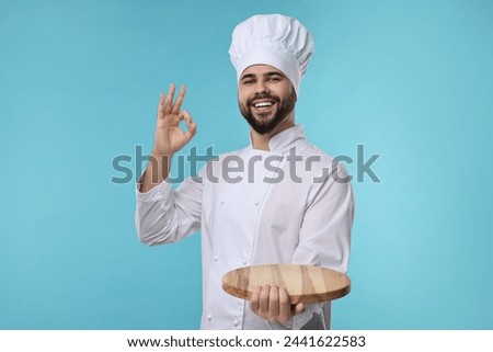 Happy young chef in uniform holding wooden board and showing ok gesture on light blue background