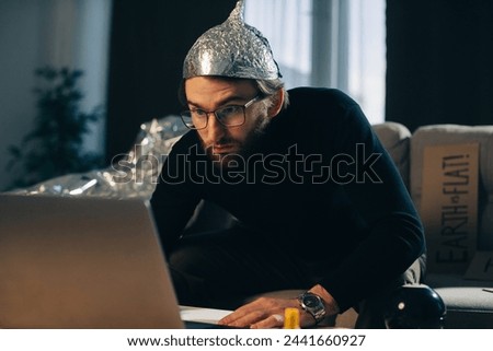 Conspiracy theory. A man in a tinfoil hat looks for signs while watching a video on a laptop.