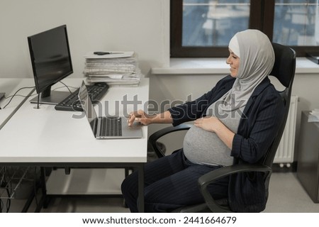 Pregnant Caucasian woman in hijab working at a computer in the office. 