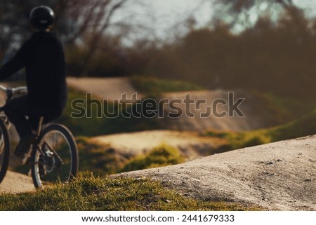 dirt bike teen coasting on a dirt jump track towards start ramp in bright sunlight. selective focus on dirt jump in foreground, rider blanked out blurry in background. Extreme sport concept