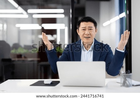 Surprised korean man spreading arms while sitting in front of personal laptop at desk in well-lit office. Evil male solving problems with help of modern technology indoors. Royalty-Free Stock Photo #2441716741