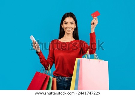 Happy woman holding multiple shopping bags, credit card and smartphone in hands, cheerful female enjoying making purchases, standing on blue background
