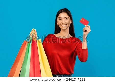 Happy young woman holding shopping bags in one hand and credit card in the other, smiling female enjoying making purchases, standing on blue studio background, free space