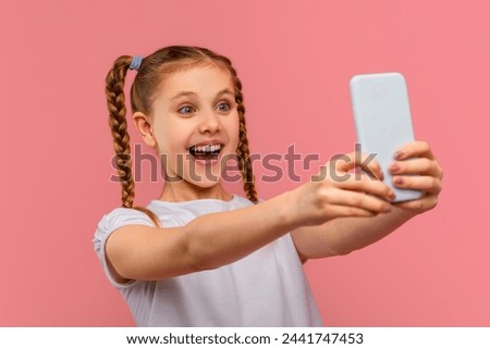 An excited young girl takes a selfie with her smartphone, looking thrilled on a pink background