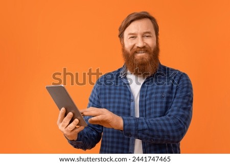 Redhaired bearded businessman in casual using digital tablet computer, smiling to camera while standing on orange backdrop in studio, portrait of man websurfing on gadget Royalty-Free Stock Photo #2441747465