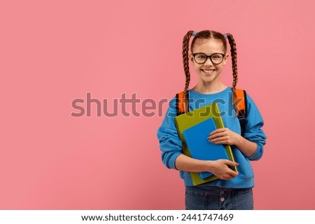 A happy schoolgirl with braids holding blue books and wearing a backpack smiles at the camera