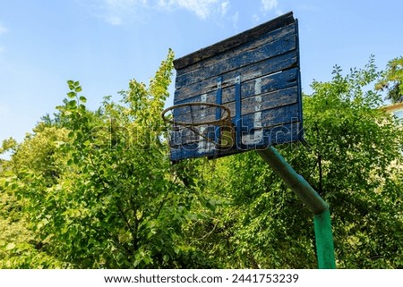 A basketball hoop is in a park with trees around it. The hoop is old and rusted, and the net is torn. The park is empty, and the sky is clear