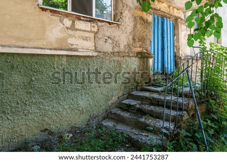 A blue door with a blue trim sits in front of a building with a green wall. The door is open and the stairs leading up to it are rusted. The scene is somewhat eerie and abandoned