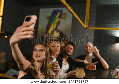 Pretty young woman with glass of alcoholic drink in hand takes photo on smartphone of friends. Joyful young people drink cocktails discussing news
