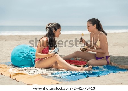 Latin young female friends enjoying beach picnic eating watermelon and taking pictures during holidays on a sandy beach