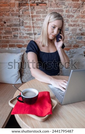 This image captures a young Caucasian woman with blonde hair engaged in a busy workday at a cafe. She's multitasking efficiently, with one hand on her laptop keyboard and the other holding a