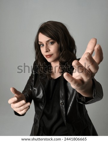 close up portrait of beautiful brunette woman wearing long black leather trench coat. Gestural hand poses, arms reaching towards camera, with wide angel perspective, Isolated on studio background