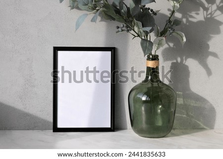 Blank picture frame mockup, green vase with tree branches on marble table, gray textured concrete wall background with natural abstract sunlight shadows, aesthetic cozy home interior design.