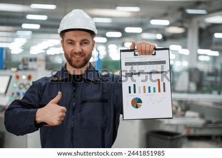 Plan with graphs in hand. Factory worker is indoors with hard hat.