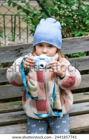 A young girl is sitting on a rustic wooden bench in a park, taking pictures with a blue camera. She is wearing a warm winter coat and a blue beanie hat. The background is a green leafy blur.