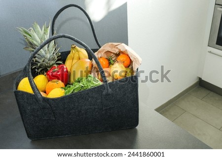 A grey eco felt bag on kitchen table with groceries, fresh vegetables and fruits. Royalty-Free Stock Photo #2441860001