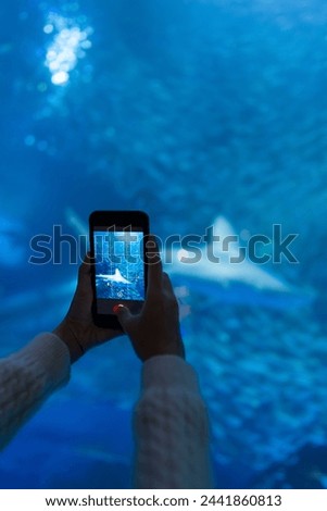 Happy Asian woman using mobile taking picture of aquatic animals in large glass tank during travel underwater zoo Aquarium. Attractive girl enjoy learning sea life at Oceanarium on holiday vacation.