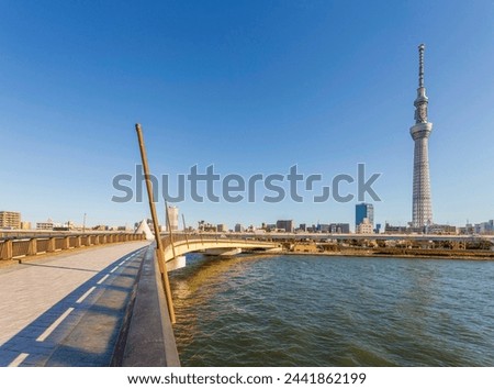 City Skyline with Tower and Riverfront Promenade, A clear day showcases a riverfront promenade leading towards a tall tower, with a bridge and city skyline in view. Royalty-Free Stock Photo #2441862199