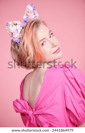 A cute blonde teenage girl with a short haircut poses in a pink dress and a lovely headband with floral kitty ears. Pink background. Kids and teenage fashion. Spring-summer look.