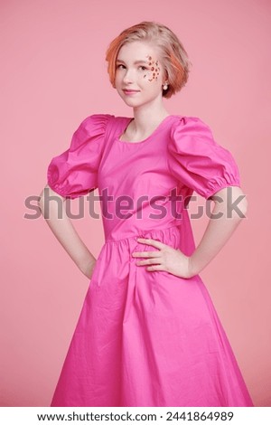 Teenage and kids Fashion. A modern teen girl with a short blonde hair with orange streaks poses with flower stickers on her face, wearing a pink dress. Pink background. Gentle spring-summer look.