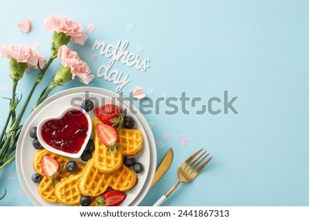 Mother's Day breakfast table: top view heart waffles, strawberries, blueberries, sweet syrup, cutlery, carnations, note "mothers day," on a light blue surface, area for message Royalty-Free Stock Photo #2441867313
