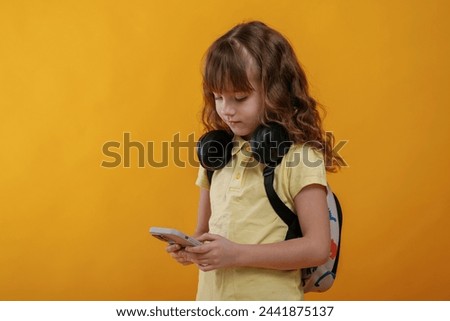 Holding smartphone, headphones on the neck. Cute little girl is against yellow background.