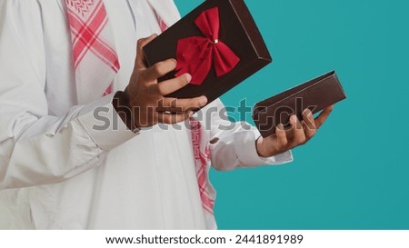 Islamic man opening giftbox with bow, receiving amazing present on his birthday. Middle eastern person feeling pleased about gift wrapped with ribbon, holiday package in studio.