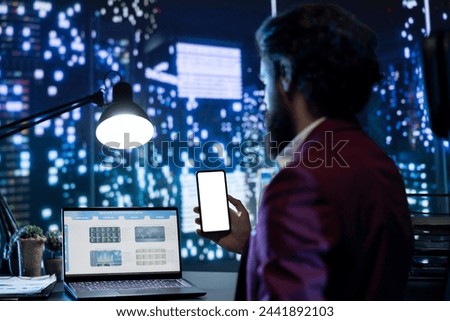 Financial advisor working on boosting earnings at night, holding phone with white screen mockup in opulent office. Company CEO analyzing monthly transactions, using blank copyspace layout. Royalty-Free Stock Photo #2441892103