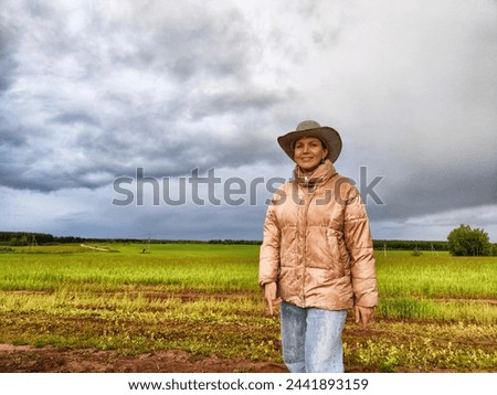 An adult girl looking like a cowboy in a hat in a field and with a stormy sky with clouds takes pictures of a rainbow and takes selfie in the rain. Woman having fun outdoors on rural and rustic nature