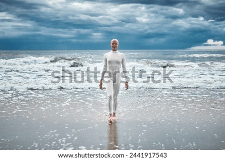 Brave hairless girl with alopecia in white futuristic suit comes out of cold sea on sandy beach, metaphoric performance of bald female artist about overcoming challenges of life and self acceptance