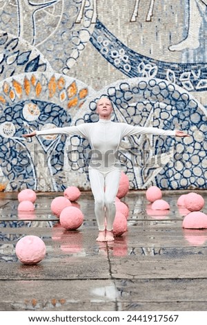 Young hairless girl ballerina with alopecia in white futuristic suit stands outdoor spreading arms to sides among pink spheres on abstract mosaic Soviet background, symbolizes self expression