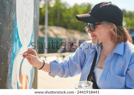 Young female painter in black cap and sunglasses passionately paints picture with paintbrush for outdoor street exhibition, female artist engrossed in creating vibrant artwork at bright sunny day