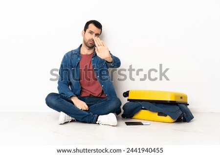 Caucasian handsome man with a suitcase full of clothes sitting on the floor at indoors making stop gesture