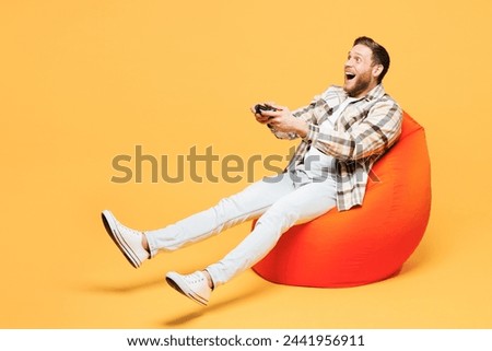 Full body young Caucasian man he wear brown shirt casual clothes sit in bag chair hold in hand play pc game with joystick console isolated on plain yellow orange background studio. Lifestyle concept