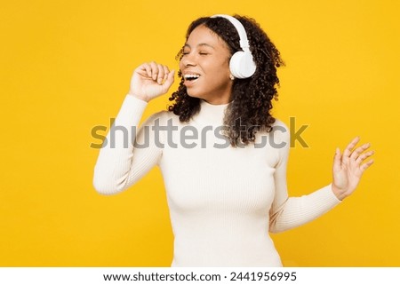 Little kid teen girl of African American ethnicity wear white casual clothes listen music in headphones sing song in microphone isolated on plain yellow background studio. Childhood lifestyle concept