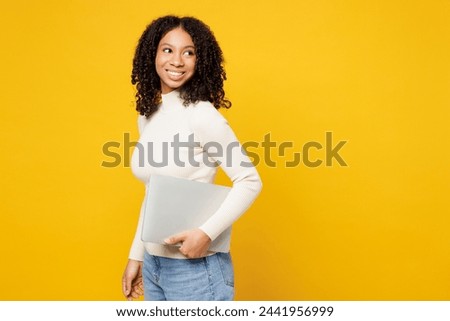 Side view little IT kid teen girl of African American ethnicity wears white casual clothes hold laptop pc computer look aside isolated on plain yellow background studio. Childhood lifestyle concept