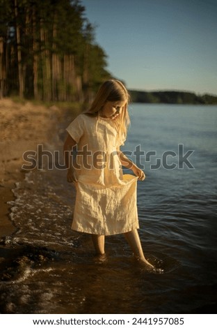 a seven-year-old fair-haired girl in a linen dress stands at the edge of the water, childlike curiosity and spontaneity, summer activity on the water, a healthy lifestyle, a cheerful childhood Royalty-Free Stock Photo #2441957085
