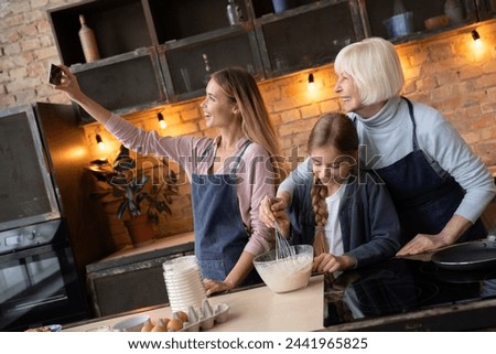 Happy young mother taking selfie with her daughter and grandmother in kitchen. Grandmother and little girl making food in kitchen while mother taking self portrait with mobile phone