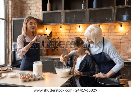 Happy young mother taking picture on mobile phone of her daughter and grandmother in kitchen. Grandmother and little girl making food in kitchen while mother taking picture with mobile phone