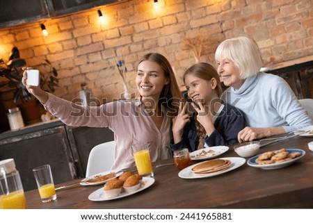 Happy young mother taking selfie with her daughter and grandmother in kitchen. Grandmother and little girl having fun in kitchen while mother taking self portrait with mobile phone