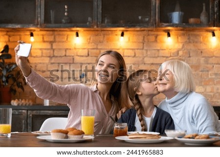 Front shot of smiling young woman taking photo selfie of her kissing grandmother daughter while having breakfast on modern kitchen. Friendly close family doing self shoot for memories