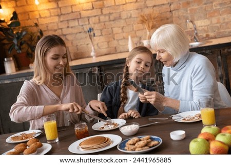 Horizontal shot of happy family at the kitchen table dining. Smiling grandmother taking picture of the plate with pancakes that eating granddaughter. Happy family concept