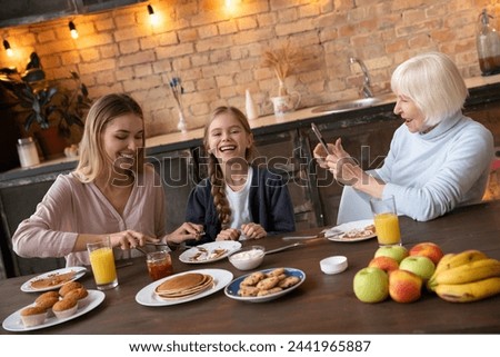 Indoor shot of laughing family at the kitchen table on breakfast. Smiling grandmother taking picture of her happy daughter and granddaughter eating bakery. Happy family concept