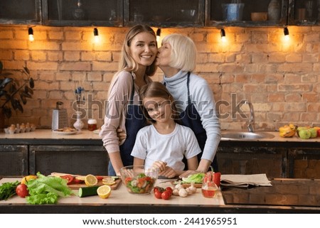 Cheerful senior woman kissing her daughter in home kitchen. Cute girl standing near them and looking in camera. Happy family portrait of elderly woman with daughter and grandchild during cooking